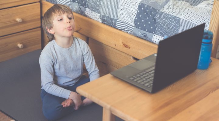 Young boy sits cross-legged on a mat in a bedroom, looking at an open laptop on a wooden table beside a bed with a patchwork quilt. - Кому підходить онлайн логопед: переваги