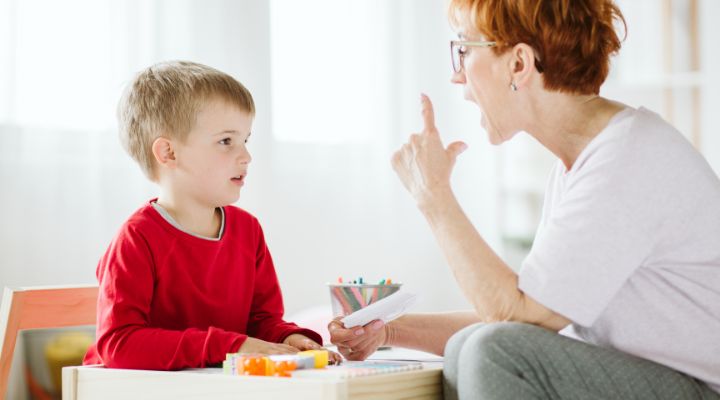 Grandmother and young boy sit at a play table; she points a finger as she speaks, signaling a scolding or calm instruction moment during playtime. - Що таке запуск мовлення