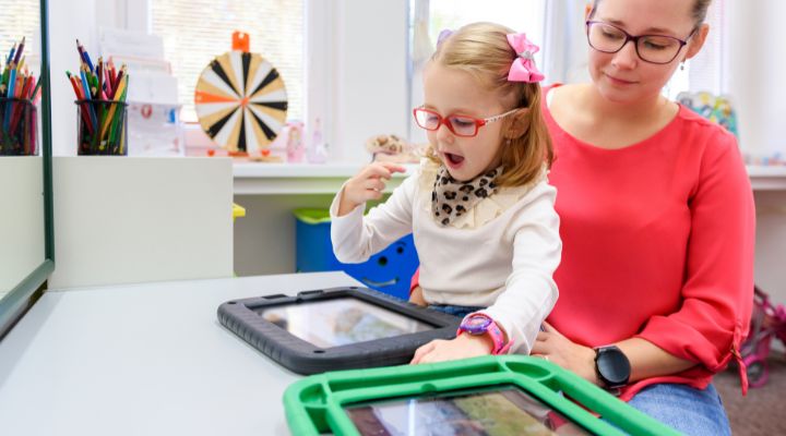 Young girl with pink glasses and a scarf uses a tablet while a woman in a red top supervises in a bright classroom. - ЗНМ 1, 2, 3 рівень: пояснення для батьків