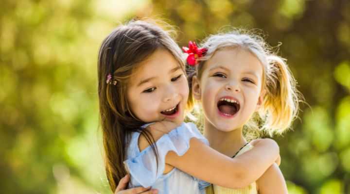 Two young girls hug and laugh outdoors on a sunny day in a park setting. - Закрита й відкрита ринолалія: різниця